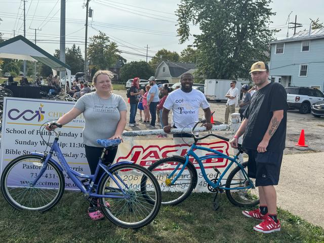 Two neighbors with their new bikes at the bike giveaway, with Pastor Harris behind them